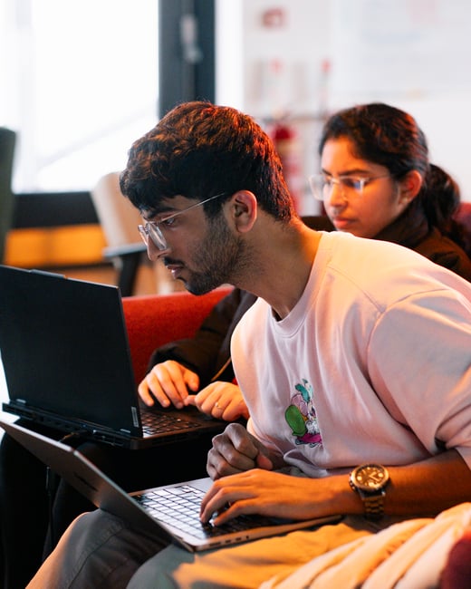 A close-up shot of a young man with glasses and a pink sweatshirt focused on his laptop, while others work on their laptops in the background.