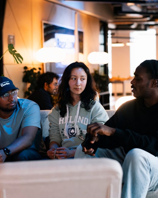 A group of young adults are sitting on a couch in a warmly lit, modern office lounge, engaged in a conversation.