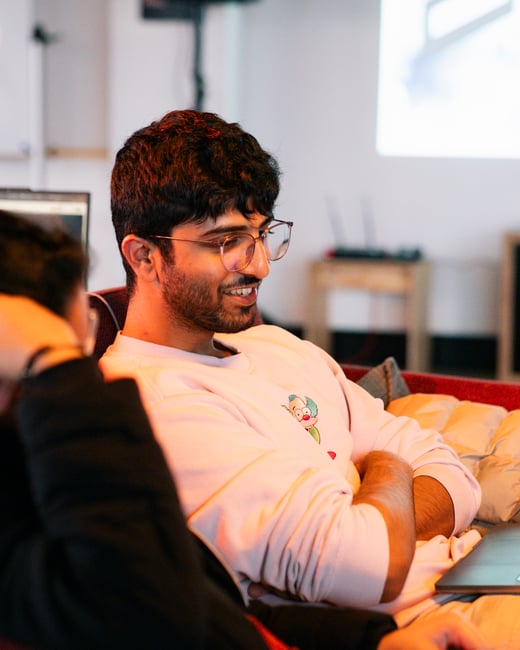 A close-up photograph of a smiling young man with glasses and a pink sweatshirt looking at a laptop screen in an indoor setting.