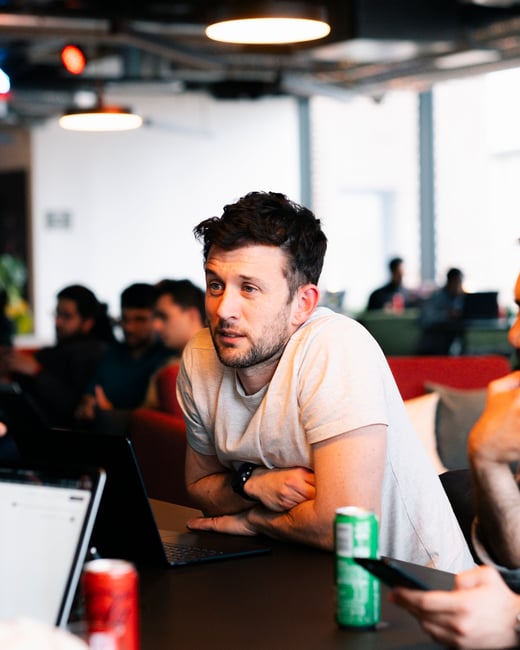 A man in a light grey t-shirt sits at a table in a modern office or coworking space, listening intently during a discussion with laptops and drinks nearby.
