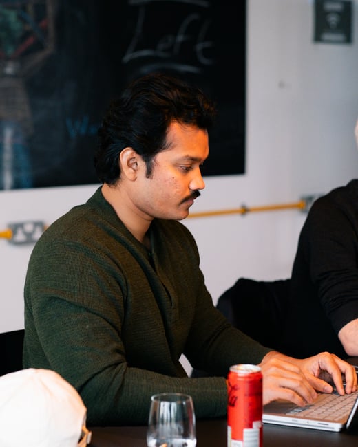 A man with dark hair and a mustache, wearing a green long-sleeve shirt, is focused on typing on a laptop at a table with a red beverage can in front of him.