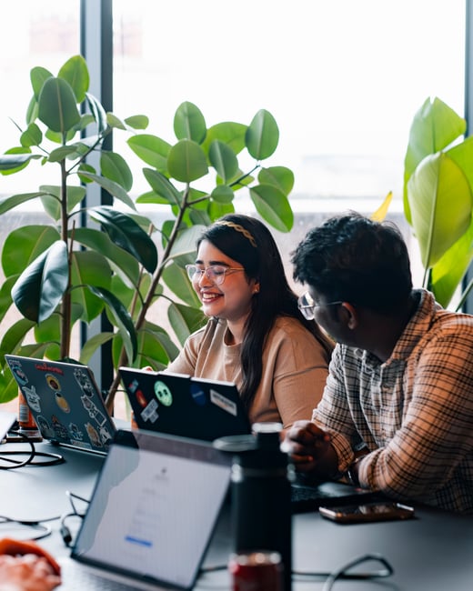 A young woman with glasses and a headband smiles while looking at a laptop, sitting next to a young man in a plaid shirt. They are in a bright indoor space with large green plants in the background.
