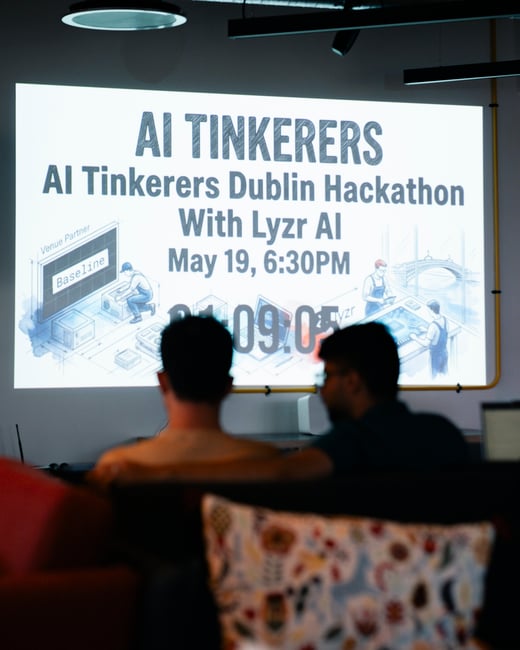 A photograph showing two individuals from behind, seated in a dimly lit room and looking at a large projection screen displaying details for the AI Tinkerers Dublin Hackathon.