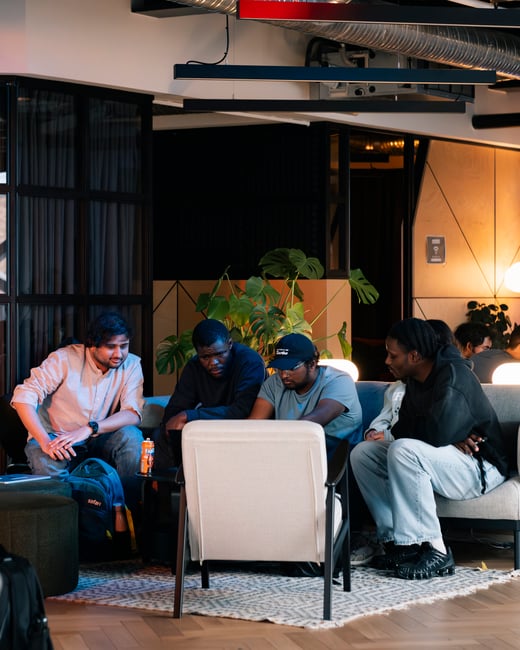 A group of young colleagues are gathered in a modern, cozy office lounge, focused on a laptop screen while sitting on a sofa and armchairs.