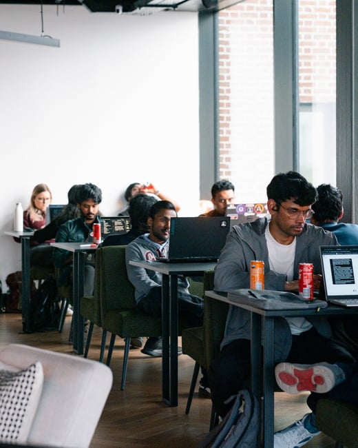 A group of young adults are sitting at individual desks in a modern, well-lit co-working space or office, focused on their laptops.