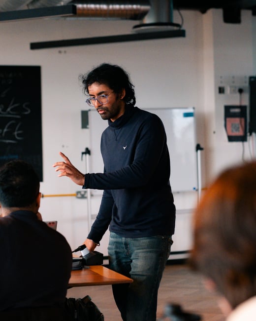 A man wearing glasses and a navy blue turtleneck is standing and gesturing while presenting to an audience in an indoor setting.