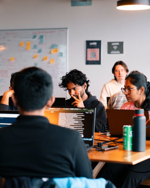 A group of people are gathered around a wooden table in an office or workshop setting, focused on their laptops with a whiteboard containing sticky notes in the background.