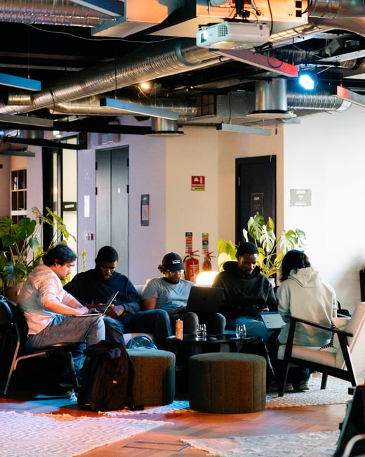 A group of five people are sitting in a modern office lounge area, focused on their laptops and phones under industrial-style ceiling pipes and warm lighting.