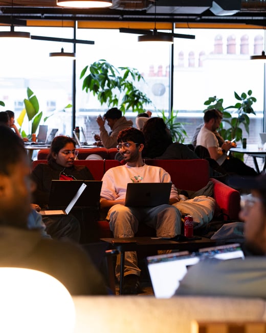 A group of people working on laptops inside a modern, warm-toned co-working space or lounge, with a man and a woman collaborating on a red sofa in the center.