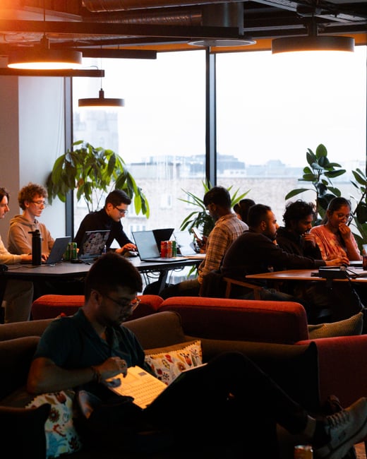 A group of people working on laptops in a modern, well-lit co-working space or office, with one person in the foreground reading on a couch.