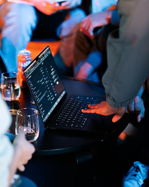 A close-up shot of a person's hand typing on a laptop keyboard at a small table, surrounded by others in a dimly lit room with blue and orange ambient lighting.
