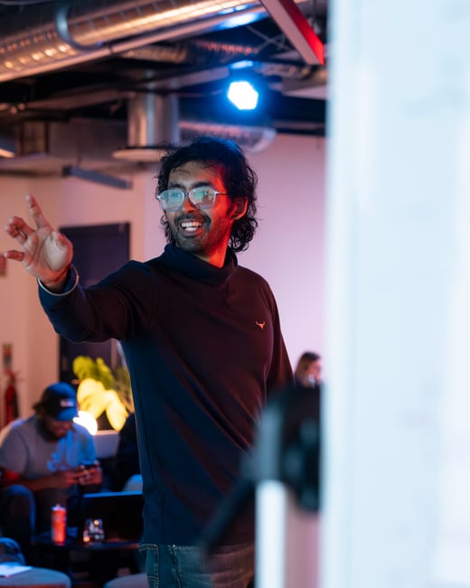 A photograph of a man with glasses and a dark turtleneck sweater smiling and gesturing with his hand during a presentation or discussion in an indoor setting with colorful lighting.