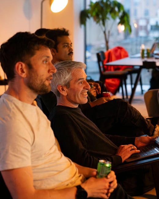A candid photograph of several men sitting indoors, focused on something off-camera, with one man smiling while using a laptop and another in the foreground holding a beverage.