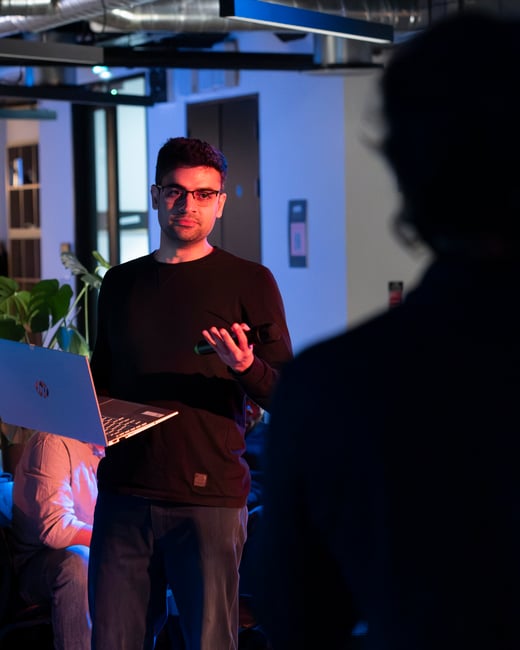 A man wearing glasses and a black long-sleeved shirt stands presenting to an audience, holding a laptop in one hand and a microphone in the other under dramatic red and blue lighting.