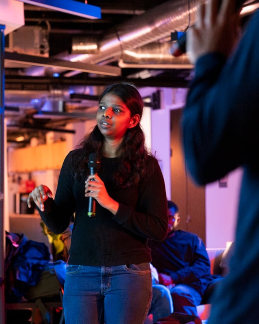 A photograph of a young woman speaking into a microphone during an event, with colorful blue and purple ambient lighting in an indoor setting.