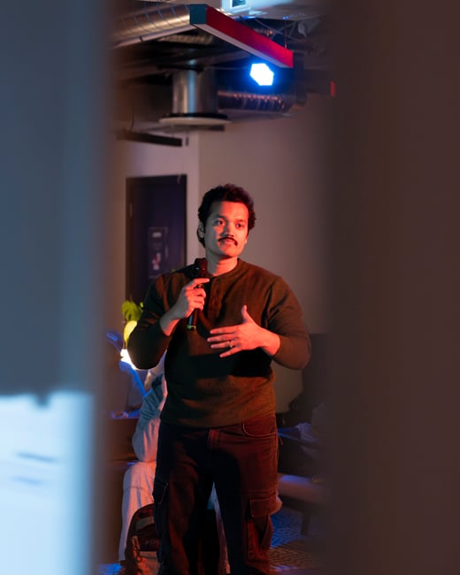A photograph of a man with a mustache speaking into a microphone, illuminated by dramatic red and blue lighting, framed between two blurred foreground pillars.