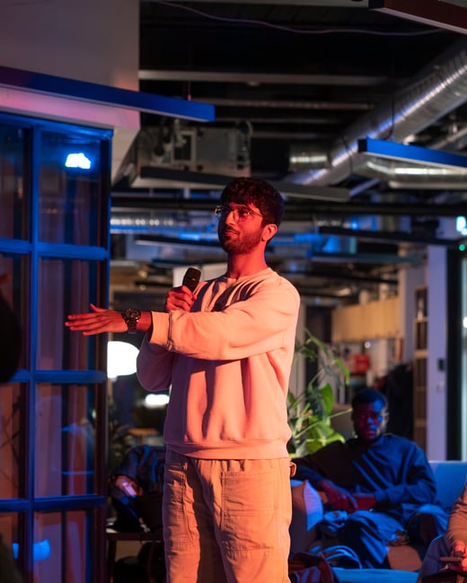 A young man wearing glasses and a sweatshirt speaks into a microphone and gestures with his hand during an indoor presentation, illuminated by blue and warm pink lighting.