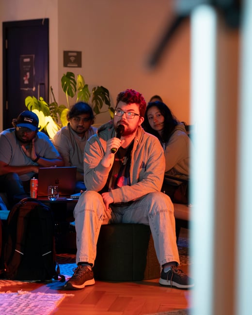 A man with glasses and a grey hoodie sits on a stool, holding a microphone and speaking, while other audience members listen behind him in a warmly lit indoor setting.