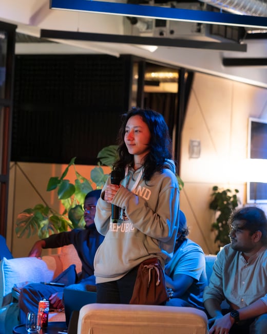 A young woman stands holding a microphone, speaking or preparing to speak at an indoor gathering, while several people sit on a couch behind her under warm and cool accent lighting.