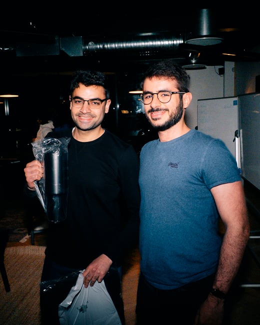 Two men standing indoors, smiling at the camera. One man on the left is holding a packaged black thermos and a plastic bag, while the man on the right wears glasses and a blue t-shirt.