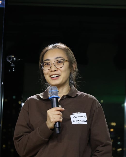 A woman wearing glasses and a brown collared shirt is speaking into a microphone, smiling brightly during an indoor evening event. She has a handwritten name tag identifying her as Aurora Li from Google Cloud.