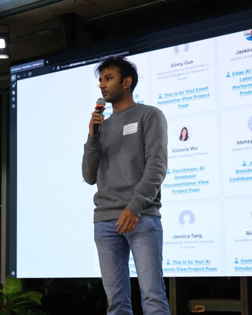 A photograph captures a young man standing on a wooden stage, speaking into a microphone while wearing a grey sweatshirt and blue jeans, with a large illuminated screen behind him displaying presenter profiles and project details.