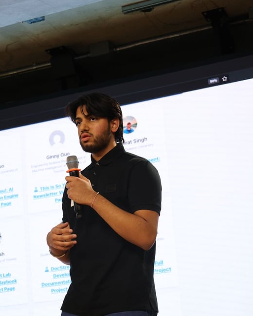 A young man wearing a black polo shirt and jeans is speaking into a microphone while standing in front of a large projection screen displaying names and project information. The setting appears to be an indoor event or conference.