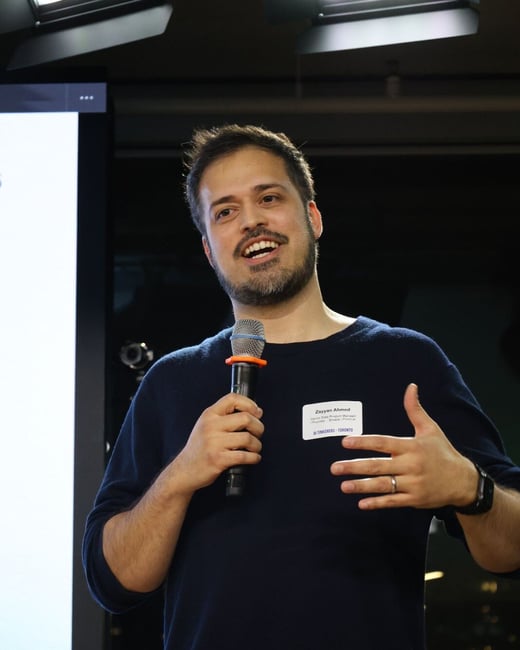 A man identified by his name tag as Zayyan Ahmed is giving a presentation or talk indoors, holding a microphone and gesturing with his hand while smiling. He is wearing a dark blue sweater, and a bright white screen is partially visible behind him on the left.