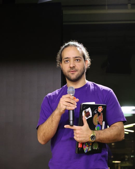 A man wearing a bright purple t-shirt stands on a stage or dark setting, holding a microphone in one hand and a black laptop or notebook covered in stickers in the other.