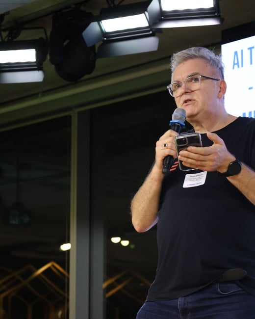 A candid photograph captures a middle-aged man with gray hair and glasses speaking into a microphone while holding a smartphone during an indoor presentation or conference event. He is wearing a black t-shirt and dark jeans, standing under bright stage lighting.