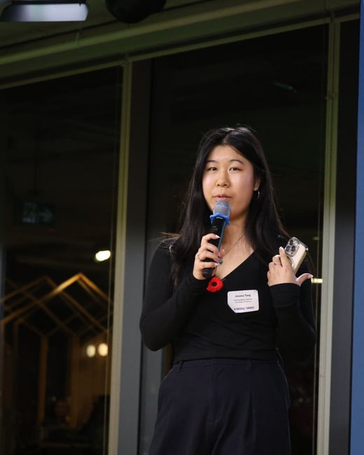 A professional photograph captures a young woman speaking into a microphone at an indoor event, wearing a dark outfit and a poppy pin on her chest.