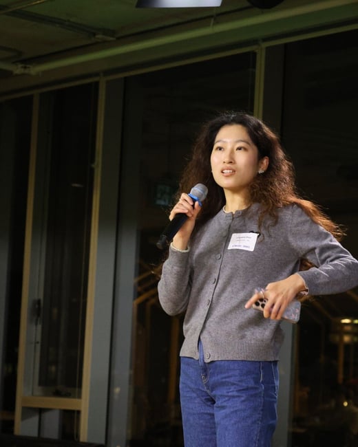 A photograph captures a woman, identified by a name tag as Catherine Chen, standing indoors and speaking into a microphone while holding a smartphone, likely during a professional presentation or event.