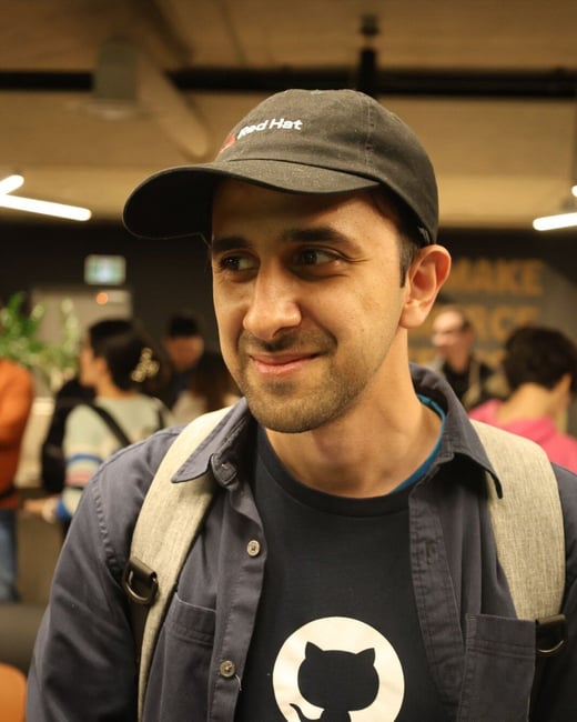 A candid, close-up photograph of a smiling man wearing a Red Hat baseball cap, a dark shirt featuring the GitHub logo, and a name badge, standing indoors at what appears to be a tech event.