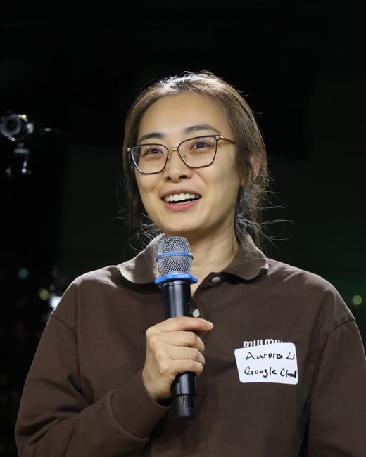 A close-up photograph captures a woman wearing glasses and a brown polo shirt, holding a microphone and smiling while speaking at an event. She has a handwritten name tag pinned to her shirt.