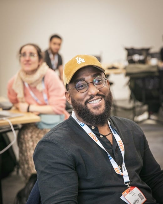 A candid portrait photograph of a smiling man wearing a black shirt, glasses, and a tan baseball cap, sitting in an audience at an indoor professional event, with other attendees blurred in the background.