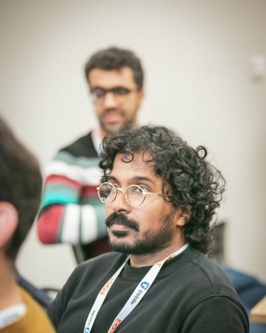 A candid close-up photo focuses on a man with curly black hair, a beard, and round gold glasses, wearing a black sweatshirt and a conference lanyard, looking intently off-camera to the side. Another man in a striped sweater stands blurred in the background.