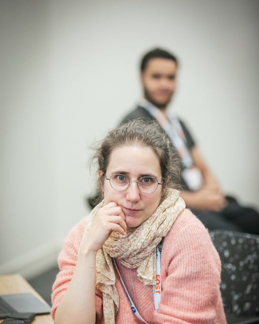 A candid, close-up photograph captures a woman wearing glasses, a pink sweater, and a scarf, seated at a table with a laptop, looking directly at the camera while a blurred man sits in the background.