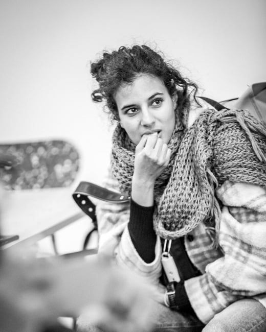 A striking black and white portrait captures a young woman with curly hair, wearing a thick knitted scarf and plaid jacket, seated indoors while looking away contemplatively, with her fingers near her mouth. The image uses a shallow depth of field, blurring a second person in the immediate foreground.