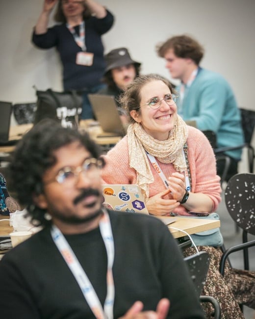 A candid photograph captures several attendees at an indoor conference or workshop, with the focus on a smiling woman wearing glasses and a pink sweater in the mid-ground, while a man in the foreground is blurred.