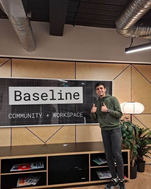 A young man, wearing an olive green hoodie and dark jeans, stands smiling and giving a double thumbs-up in the lobby of a modern workspace called 'Baseline Community + Workspace'.