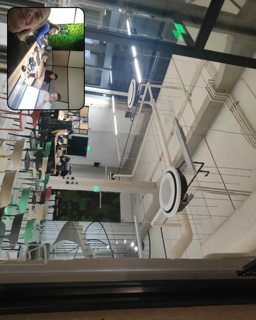 A digital photograph in the BeReal format, showing a high-ceilinged, modern office or communal space with exposed ductwork, overlaid with a small inset selfie of four people sitting around a desk with laptops.