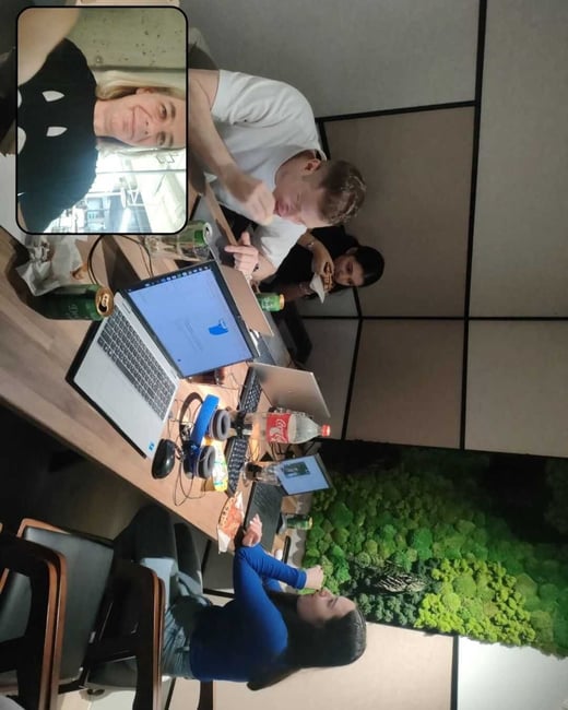 A rotated candid photograph, likely a BeReal post, showing four people gathered around a conference or work table with laptops, food, and drinks, featuring a selfie inset in the upper left corner. The indoor setting includes a decorative green moss wall in the background.