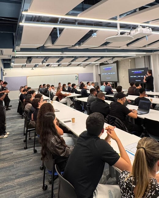 An indoor photograph capturing a presentation or lecture setting, where a speaker stands at the front of a classroom-style room addressing a large seated audience.