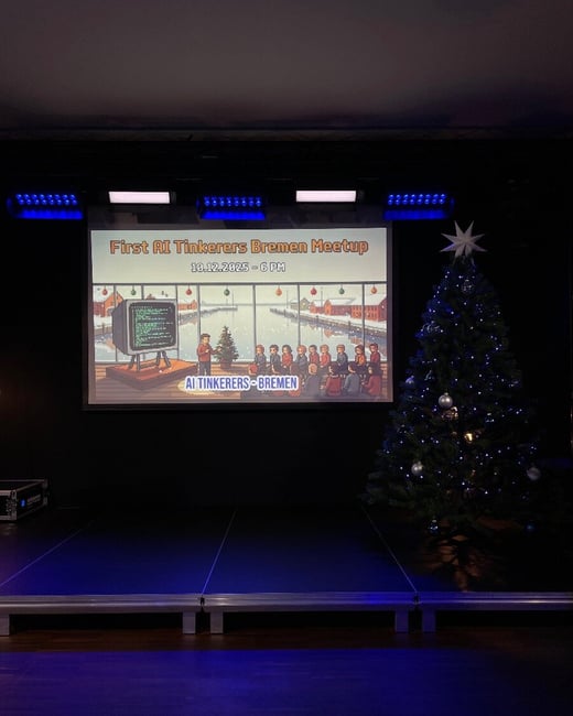 A dark, indoor photograph showing an event stage setup with a large projection screen displaying an announcement slide for the 'First AI Tinkerers Bremen Meetup' next to a decorated Christmas tree.