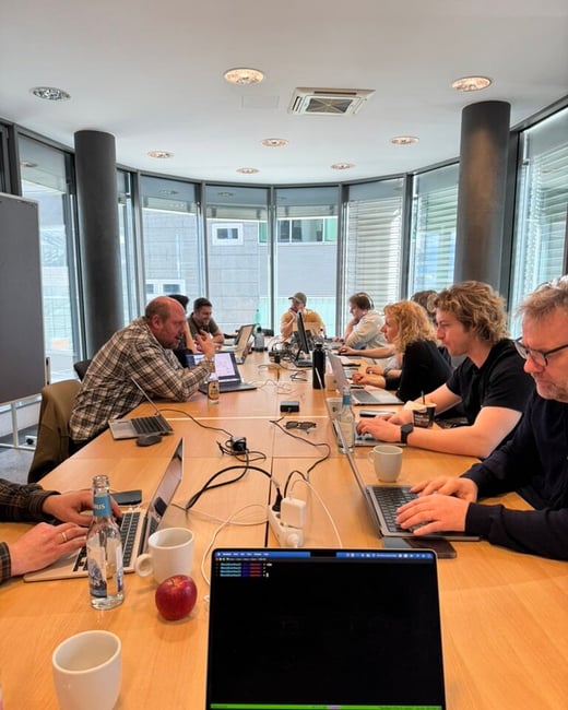 A group of people are seated around a long wooden conference table in a modern, well-lit office, focused on their laptops and engaged in a collaborative work session.