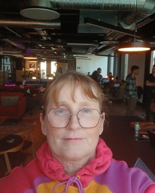 A close-up selfie of a woman with red hair and glasses, wearing a vibrant multi-colored hoodie in a modern indoor workspace.