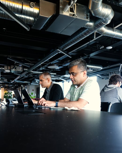 A vertical photograph showing several men working on laptops at a long black table in a modern office with an industrial-style ceiling featuring exposed pipes and ductwork.
