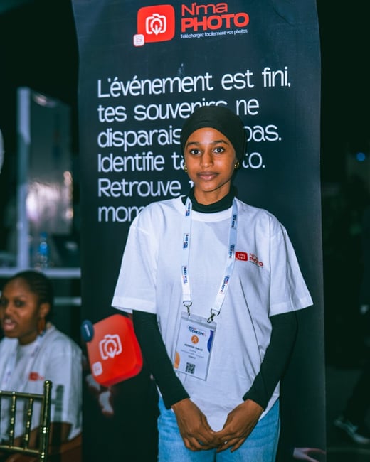 A young woman wearing a hijab and a white branded t-shirt stands in front of a promotional banner for 'N'ma PHOTO' at an indoor event.