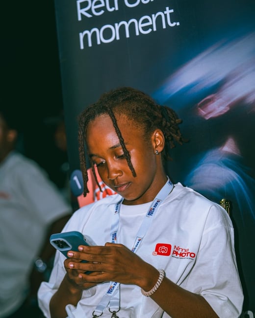 A young person with dreadlocks is focused on their smartphone while wearing a white event t-shirt and a lanyard.