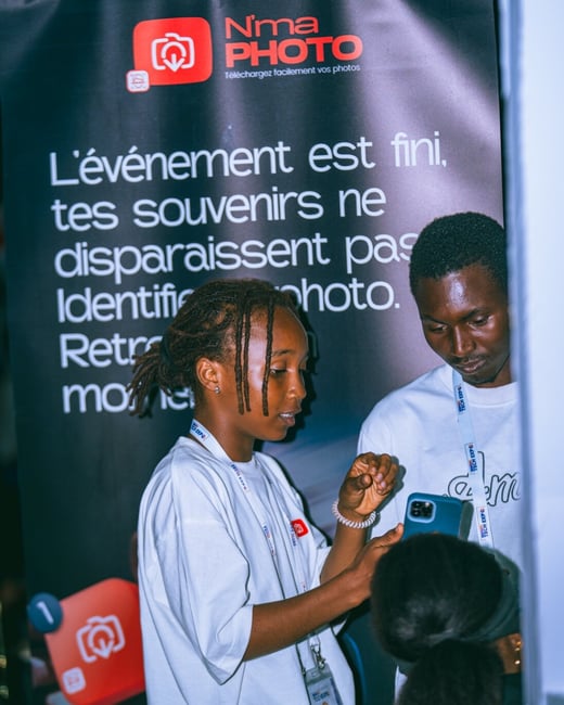 A photograph showing two young people at an event looking at a smartphone together in front of a large promotional banner.